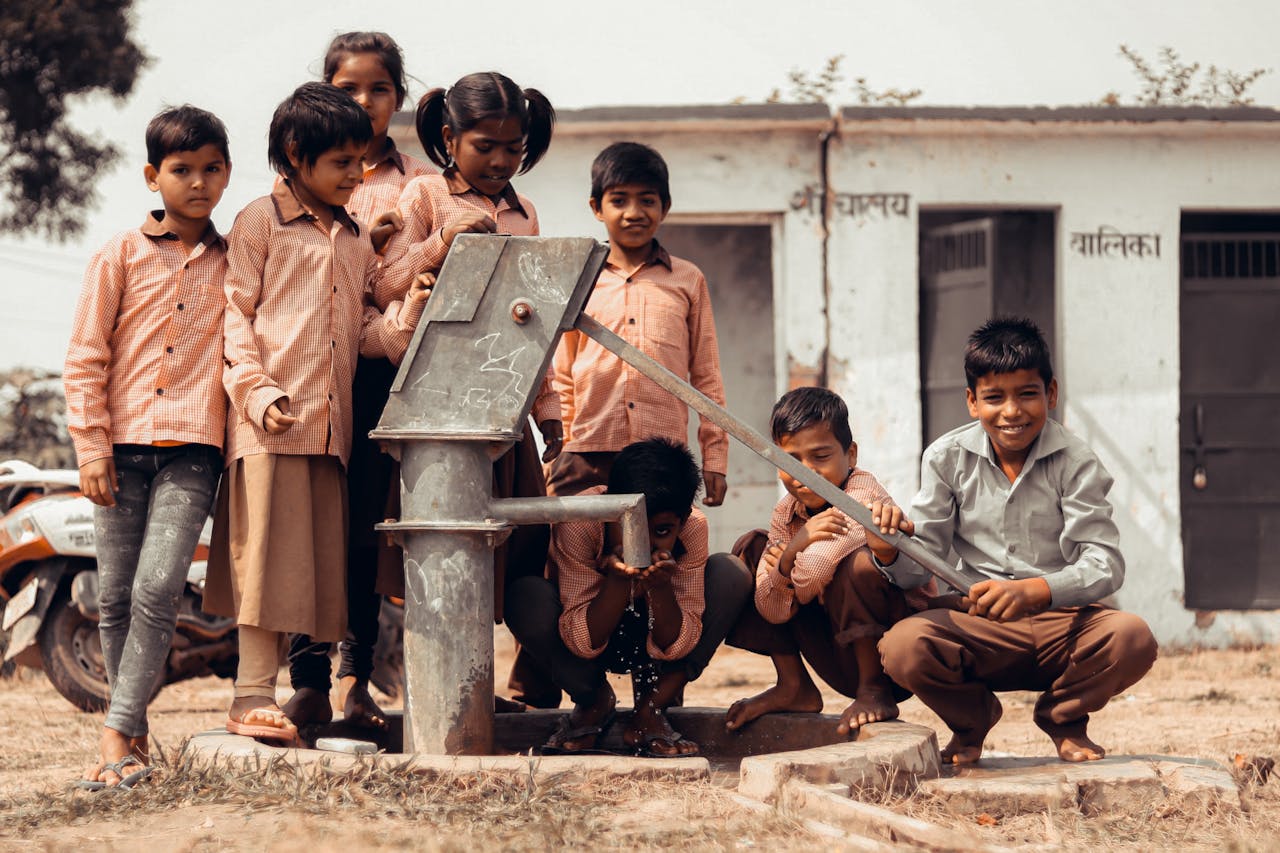A group of Indian children gather joyfully around a manual water pump outdoors.